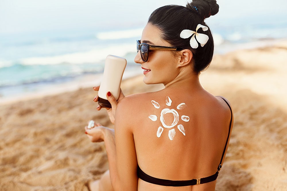 Fiatal nő vízparton, háttal, kalapban, a hátán naptejből nap rajzolva