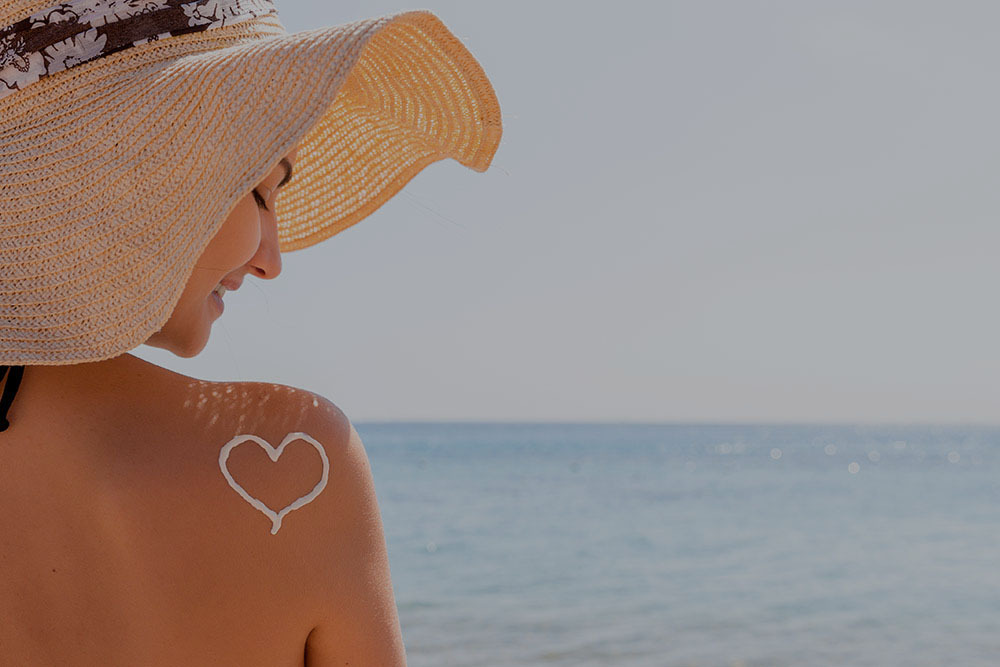 Fiatal nő vízparton, háttal, kalapban, a hátán naptejből szív rajzolva