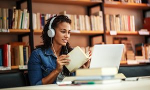 Egyetemista lány a könyvtárban tanul, kezében könyv, fején fejhallgató, az asztalon laptop
